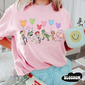 Puede incluir: Sudadera rosa claro con una ilustraci&oacute;n de dibujos animados de personajes de Toy Story sosteniendo globos con forma de Mickey Mouse. Los personajes incluyen a Woody, Buzz Lightyear, Jessie, Rex, Slinky Dog y Hamm. El texto "Buzz Lightyear" tambi&eacute;n es visible.