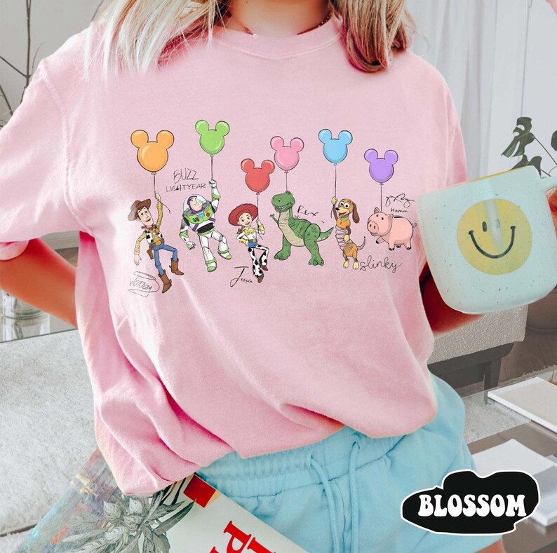 Puede incluir: Sudadera rosa claro con una ilustraci&oacute;n de dibujos animados de personajes de Toy Story sosteniendo globos con forma de Mickey Mouse. Los personajes incluyen a Woody, Buzz Lightyear, Jessie, Rex, Slinky Dog y Hamm. El texto "Buzz Lightyear" tambi&eacute;n es visible.