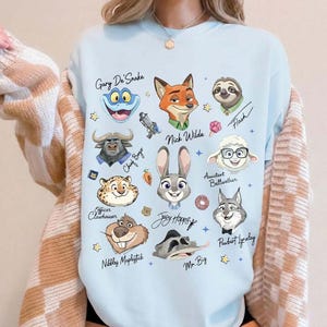 May include: Light blue sweatshirt featuring cartoon illustrations of characters from Zootopia, including Nick Wilde, Judy Hopps, and Flash. Character names are written in cursive script. The sweatshirt has a crew neck.