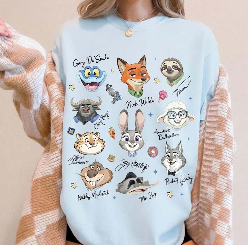 May include: Light blue sweatshirt featuring cartoon illustrations of characters from Zootopia, including Nick Wilde, Judy Hopps, and Flash. Character names are written in cursive script. The sweatshirt has a crew neck.