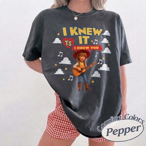 May include: A charcoal gray short-sleeve t-shirt featuring a graphic of a cowgirl playing an acoustic guitar. The text reads I KNEW IT TS I KNEW YOU, surrounded by white clouds, yellow stars, and musical notes. The shirt has a crew neckline.