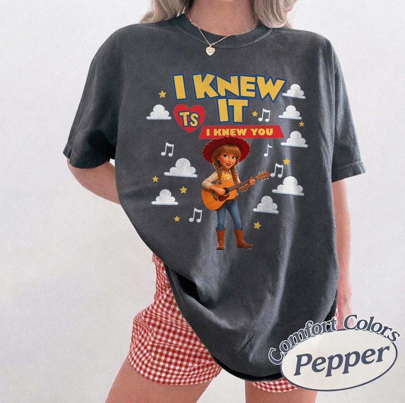 May include: A charcoal gray short-sleeve t-shirt featuring a graphic of a cowgirl playing an acoustic guitar. The text reads I KNEW IT TS I KNEW YOU, surrounded by white clouds, yellow stars, and musical notes. The shirt has a crew neckline.