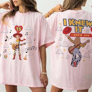 Puede incluir: Una camiseta rosa claro con un gr&aacute;fico de una vaquera tocando la guitarra en la parte delantera y una persona tocando la guitarra en la parte trasera. La espalda incluye el texto I KNEW IT I KNEW YOU y un coraz&oacute;n rojo con TS. Incluye nubes y notas.