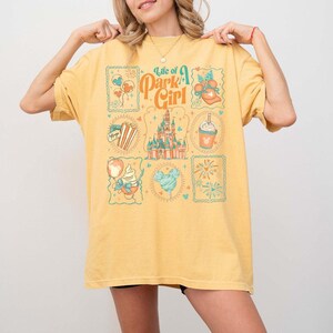 May include: Yellow t-shirt with a graphic design featuring the text "Life of a Park Girl" and various theme park-themed illustrations. The design includes images of a castle, popcorn, ice cream, and fireworks.