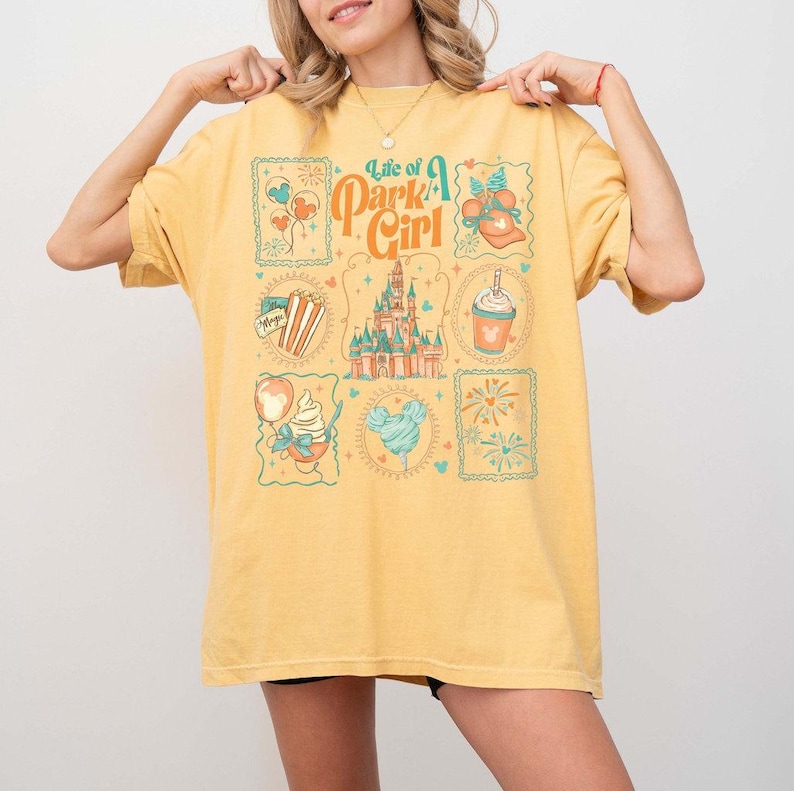 May include: Yellow t-shirt with a graphic design featuring the text "Life of a Park Girl" and various theme park-themed illustrations. The design includes images of a castle, popcorn, ice cream, and fireworks.