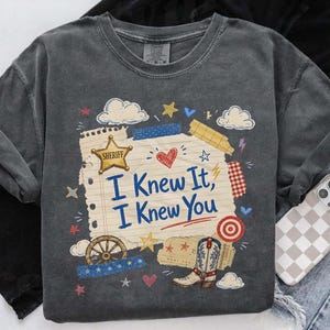 Op de afbeelding: Een antracietgrijs t-shirt met een afbeelding van een vel gelinieerd notitiepapier met de tekst "I Knew It, I Knew You" in het blauw. Het ontwerp bevat een gouden sheriffster, een cowboylaars, een wagenwiel, wolken, hartjes en sterren.