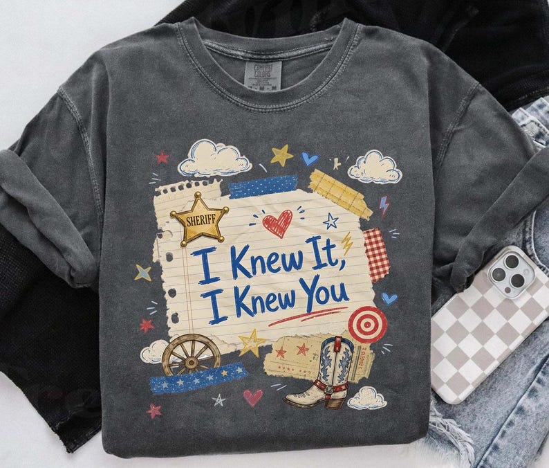 Op de afbeelding: Een antracietgrijs t-shirt met een afbeelding van een vel gelinieerd notitiepapier met de tekst "I Knew It, I Knew You" in het blauw. Het ontwerp bevat een gouden sheriffster, een cowboylaars, een wagenwiel, wolken, hartjes en sterren.