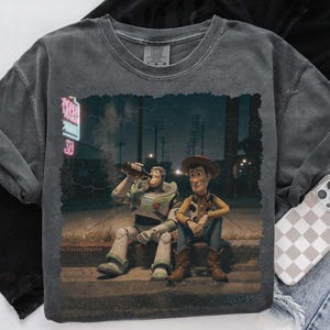 Puede incluir: Sudadera gris oscuro con cuello redondo con un gr&aacute;fico de Buzz Lightyear y Woody de Toy Story. Buzz est&aacute; bebiendo de una botella y Woody est&aacute; sentado a su lado. La imagen tiene un aspecto vintage.