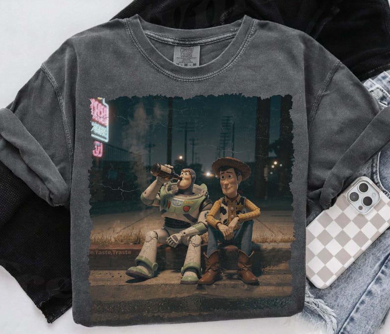 Puede incluir: Sudadera gris oscuro con cuello redondo con un gr&aacute;fico de Buzz Lightyear y Woody de Toy Story. Buzz est&aacute; bebiendo de una botella y Woody est&aacute; sentado a su lado. La imagen tiene un aspecto vintage.