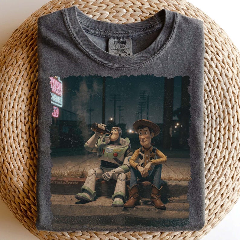 Puede incluir: Camiseta gris oscuro Comfort Colors con un gr&aacute;fico desgastado de Buzz Lightyear y Woody de Toy Story. Los personajes est&aacute;n representados sentados en una acera por la noche, con un letrero de ne&oacute;n en el fondo. La camiseta tiene cuello redondo cl&aacute;sico.