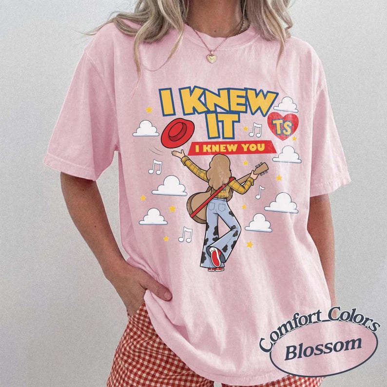 May include: A light pink short-sleeve t-shirt featuring a graphic of a person with long hair playing an acoustic guitar. The text reads I KNEW IT and I KNEW YOU, accompanied by a red hat, a red heart with TS, musical notes, and white clouds.