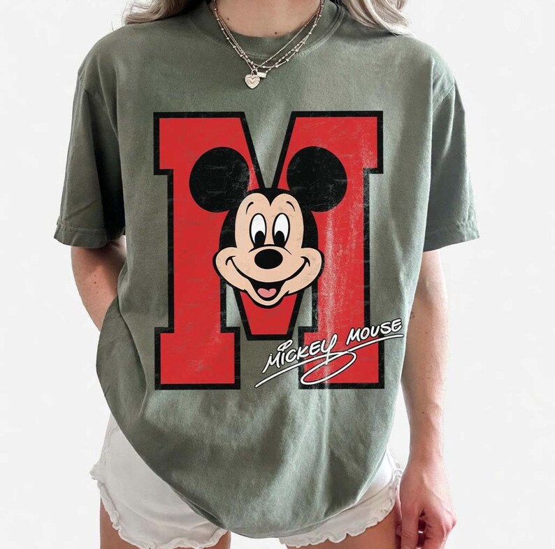 May include: Olive green t-shirt featuring a large red letter "M" with Mickey Mouse's face in the center. The words "Mickey Mouse" are written in a cursive font below the letter. The shirt has a vintage, distressed look.