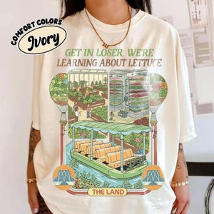 Op de afbeelding: Ivoor kleurig t-shirt met een afbeelding van een boottocht door een sla boerderij, met de tekst "GET IN LOSER, WE'RE LEARNING ABOUT LETTUCE." Het shirt heeft ook de woorden "COMFORT COLORS" en "Ivory" in een cirkel.