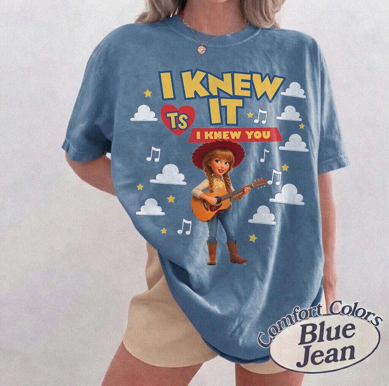 May include: A blue t-shirt featuring a graphic of a cowgirl character playing an acoustic guitar. The text reads I KNEW IT and I KNEW YOU, surrounded by white clouds, yellow stars, and musical notes. A red heart icon is also present on the shirt.