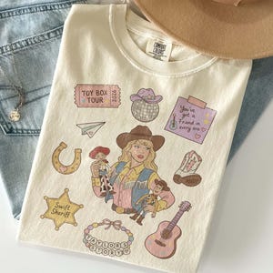 May include: A cream-colored t-shirt featuring a graphic print of a person in a cowboy hat holding two dolls. Surrounding elements include a disco ball, a guitar, a horseshoe, a star badge labeled Swift Sheriff, and a ticket reading Toy Box Tour 2026.