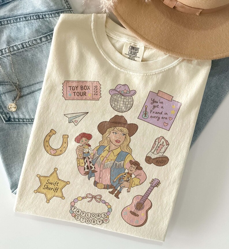 May include: A cream-colored t-shirt featuring a graphic print of a person in a cowboy hat holding two dolls. Surrounding elements include a disco ball, a guitar, a horseshoe, a star badge labeled Swift Sheriff, and a ticket reading Toy Box Tour 2026.