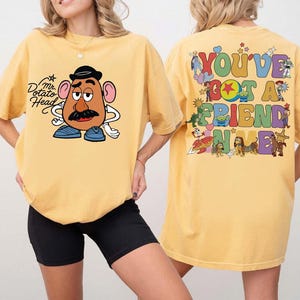 May include: A yellow t-shirt featuring a graphic of Mr. Potato Head on the front. The back displays a colorful collage of various animated characters and the text "YOU'VE GOT A FRIEND IN ME" in a bold, multi-colored, retro-style bubble font.