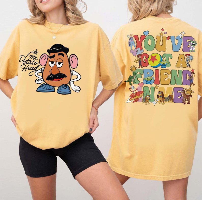May include: A yellow t-shirt featuring a graphic of Mr. Potato Head on the front. The back displays a colorful collage of various animated characters and the text "YOU'VE GOT A FRIEND IN ME" in a bold, multi-colored, retro-style bubble font.