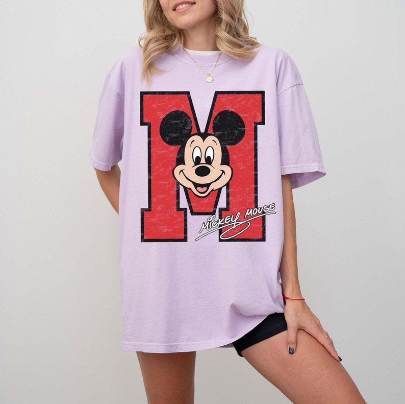 May include: A light purple t-shirt featuring a large red letter "M" with a distressed texture. Inside the "M" is a smiling Mickey Mouse graphic. The words "Mickey Mouse" are written in a stylized font below the graphic.