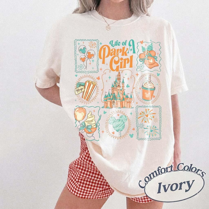 May include: Ivory-colored t-shirt with a colorful graphic design featuring the text "Life of a Park Girl" and various theme park icons. The shirt is a Comfort Colors brand.