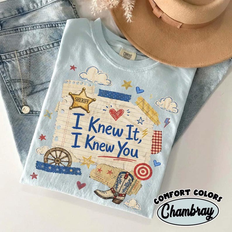 Op de afbeelding: Een lichtblauw t-shirt met een afbeelding van een notitieblokpagina met de tekst I Knew It, I Knew You. Het ontwerp bevat een gouden sheriffster, een cowboylaars, een wagenwiel, wolken, hartjes, sterren en rode en blauwe tape-accenten.