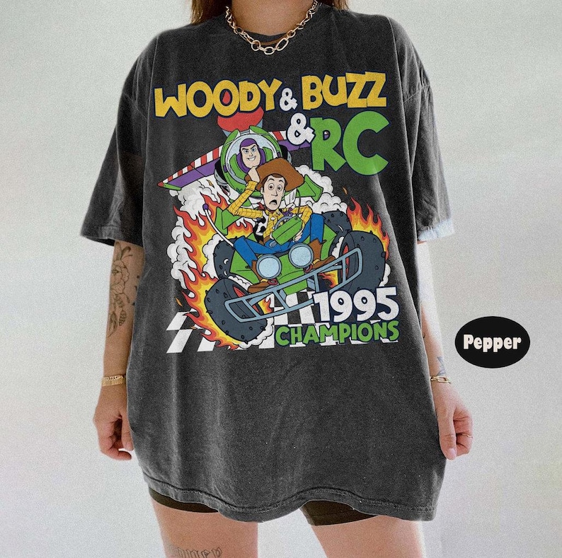 May include: Dark gray t-shirt featuring a graphic of Woody and Buzz Lightyear from Toy Story on a green RC car with flames. The text "Woody & Buzz & RC" and "1995 Champions" are also displayed.
