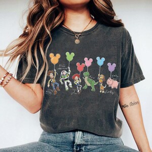 Puede incluir: Camiseta gris oscuro con personajes de Toy Story sosteniendo globos con forma de Mickey Mouse. Se representan a Woody, Buzz Lightyear, Jessie, Rex, Slinky Dog y Hamm. Los nombres de los personajes est&aacute;n escritos debajo.