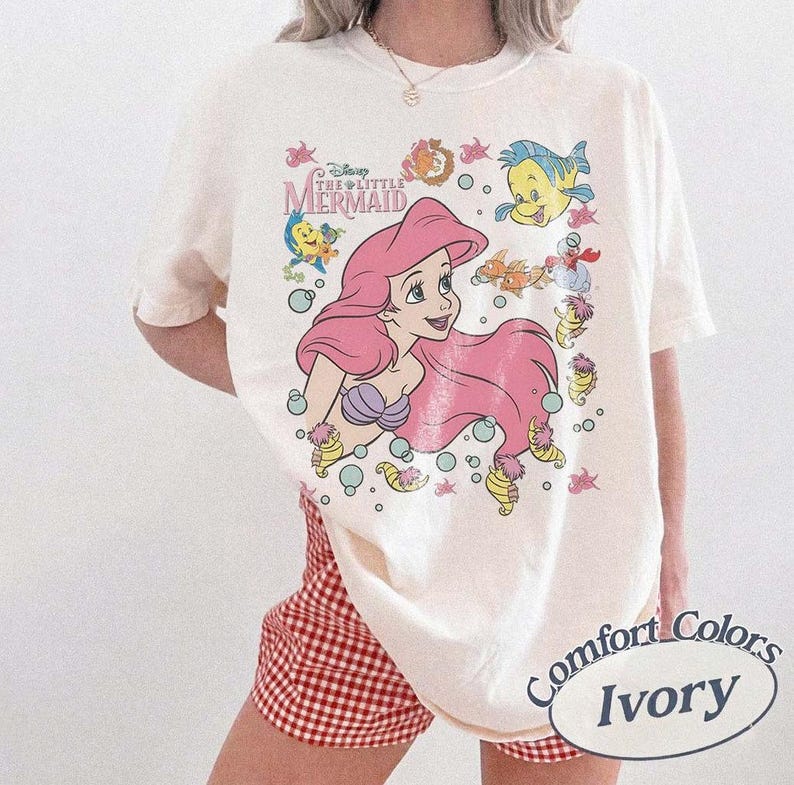 May include: Ivory-colored t-shirt featuring a graphic of Ariel from Disney's The Little Mermaid. The design includes Ariel with pink hair, sea creatures, and the film's logo. The shirt is labeled "Comfort Colors Ivory".