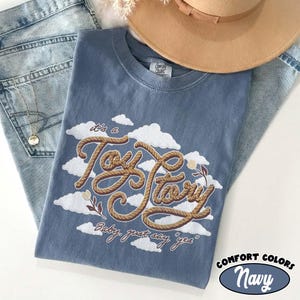 May include: A blue crew neck t-shirt featuring a graphic of white clouds and the text "it's a Toy Story" written in a brown rope-style font, with the additional text "Baby, just say 'yes'" printed below in a smaller brown cursive script.
