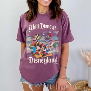 Puede incluir: Una camiseta morada con un estampado de personajes y lugares emblem&aacute;ticos de Disneyland. El texto "Walt Disney's Disneyland" est&aacute; impreso en blanco. La camiseta tiene mangas cortas y un corte holgado.