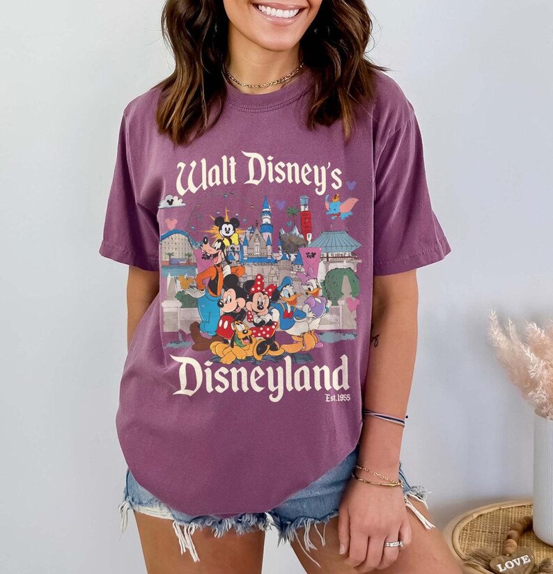 Puede incluir: Una camiseta morada con un estampado de personajes y lugares emblem&aacute;ticos de Disneyland. El texto "Walt Disney's Disneyland" est&aacute; impreso en blanco. La camiseta tiene mangas cortas y un corte holgado.