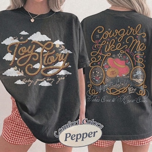 May include: A charcoal gray t-shirt featuring a front graphic with white clouds and the text "It's a Toy Story, baby, just say yes" in rope-style lettering. The back displays "Cowgirl Like Me" with a pink hat, rope, and musical notes.