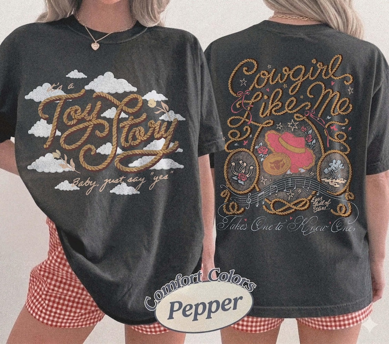 May include: A charcoal gray t-shirt featuring a front graphic with white clouds and the text "It's a Toy Story, baby, just say yes" in rope-style lettering. The back displays "Cowgirl Like Me" with a pink hat, rope, and musical notes.