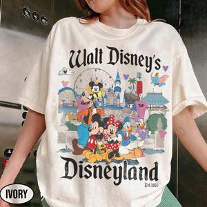 Puede incluir: Camiseta de color marfil con un gr&aacute;fico colorido de Disneyland. El dise&ntilde;o incluye a Mickey Mouse, Minnie Mouse, Donald Duck y Goofy, con las palabras "Walt Disney's Disneyland" y "Est.1955".