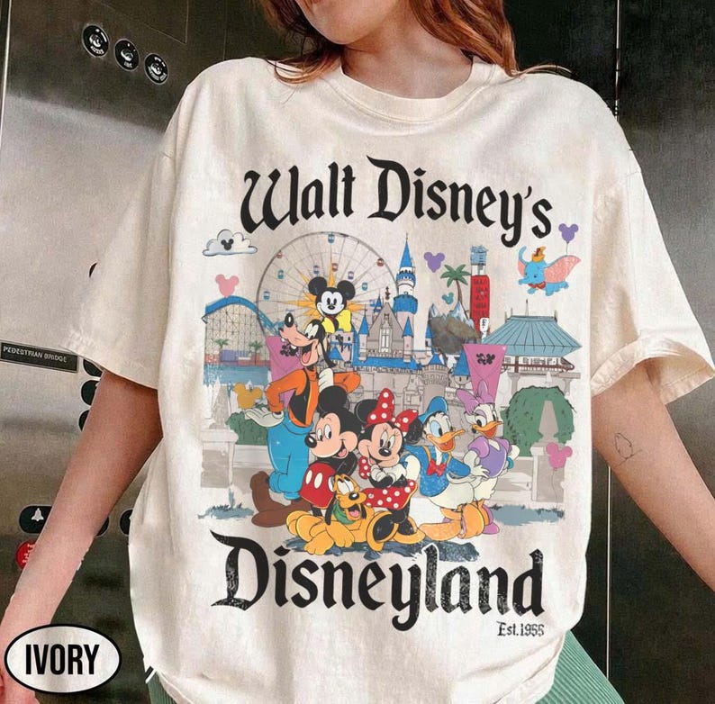 Puede incluir: Camiseta de color marfil con un gr&aacute;fico colorido de Disneyland. El dise&ntilde;o incluye a Mickey Mouse, Minnie Mouse, Donald Duck y Goofy, con las palabras "Walt Disney's Disneyland" y "Est.1955".