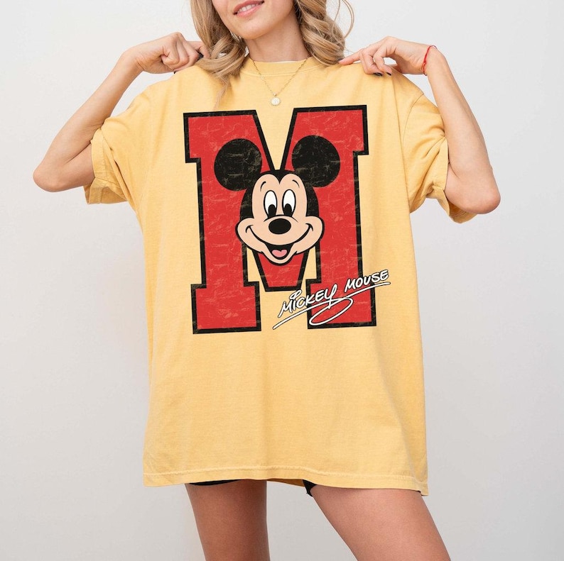 May include: Yellow t-shirt featuring a large red letter "M" with a distressed texture, incorporating a Mickey Mouse graphic. The text "Mickey Mouse" is written in a cursive font below the graphic. The shirt has short sleeves.