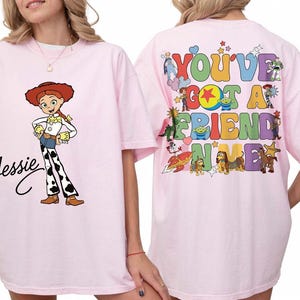 May include: A light pink t-shirt featuring a graphic of Jessie on the front with her name in black script. The back displays a colorful graphic with the text "YOU'VE GOT A FRIEND IN ME" surrounded by various animated characters and stars.