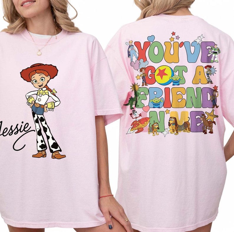 May include: A light pink t-shirt featuring a graphic of Jessie on the front with her name in black script. The back displays a colorful graphic with the text "YOU'VE GOT A FRIEND IN ME" surrounded by various animated characters and stars.