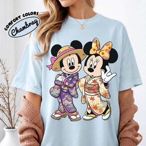 Puede incluir: Camiseta azul claro con Mickey y Minnie Mouse con trajes japoneses. Mickey lleva un sombrero de paja y un kimono morado, Minnie un lazo amarillo y un kimono crema. La camiseta tiene el texto "COMFORT COLORS Chambray".
