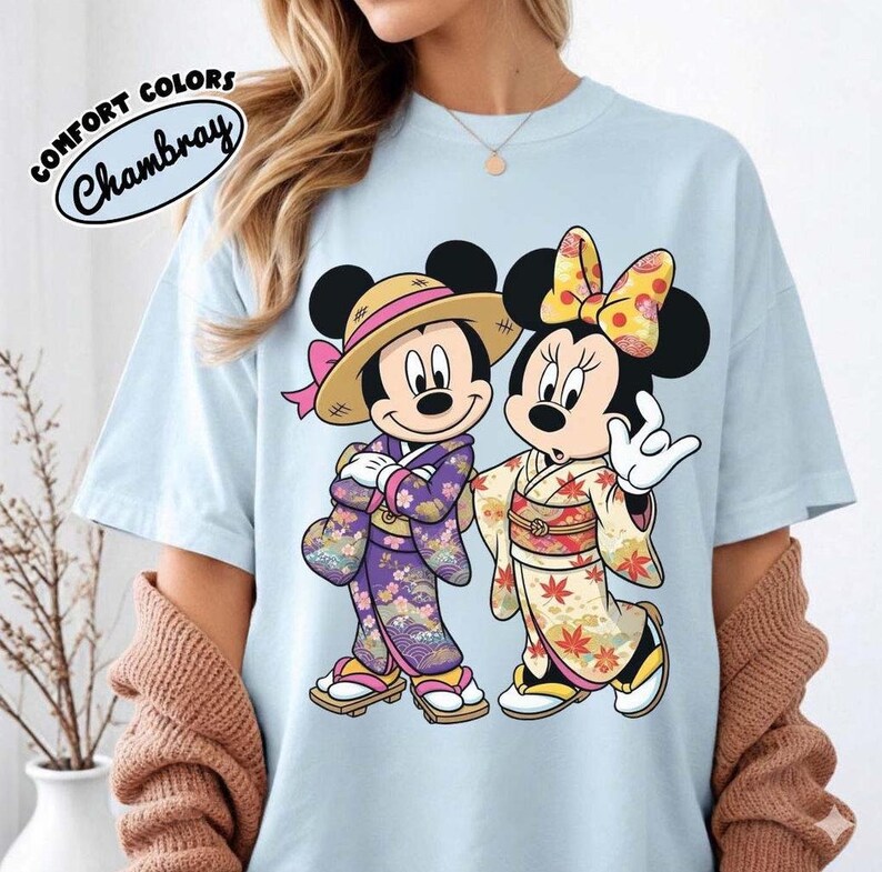 Puede incluir: Camiseta azul claro con Mickey y Minnie Mouse con trajes japoneses. Mickey lleva un sombrero de paja y un kimono morado, Minnie un lazo amarillo y un kimono crema. La camiseta tiene el texto "COMFORT COLORS Chambray".