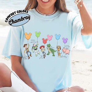 Puede incluir: Camiseta azul claro con personajes de dibujos animados sosteniendo globos con forma de Mickey Mouse. La camiseta tiene las palabras "Comfort Colors Chambray" en un dise&ntilde;o circular. Los personajes incluyen a Woody, Buzz Lightyear, Jessie, Rex, Slinky Dog y Hamm.