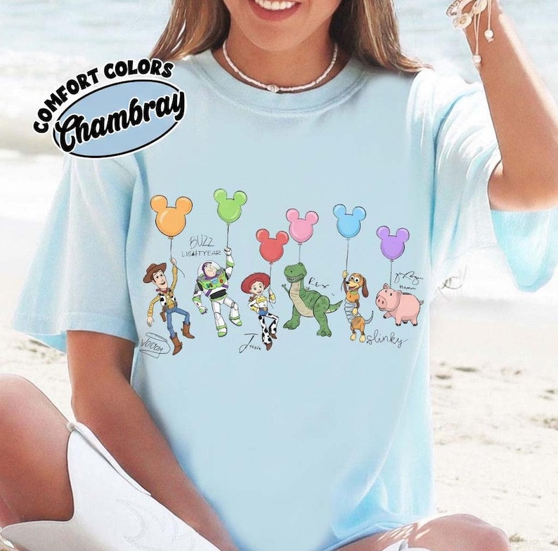 Puede incluir: Camiseta azul claro con personajes de dibujos animados sosteniendo globos con forma de Mickey Mouse. La camiseta tiene las palabras "Comfort Colors Chambray" en un dise&ntilde;o circular. Los personajes incluyen a Woody, Buzz Lightyear, Jessie, Rex, Slinky Dog y Hamm.