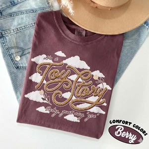 May include: A berry-colored t-shirt featuring a graphic of white clouds and the text "it's a Toy Story" written in a brown rope-style font, with the additional text "Baby, just say "yes"" printed below in a smaller white script font.