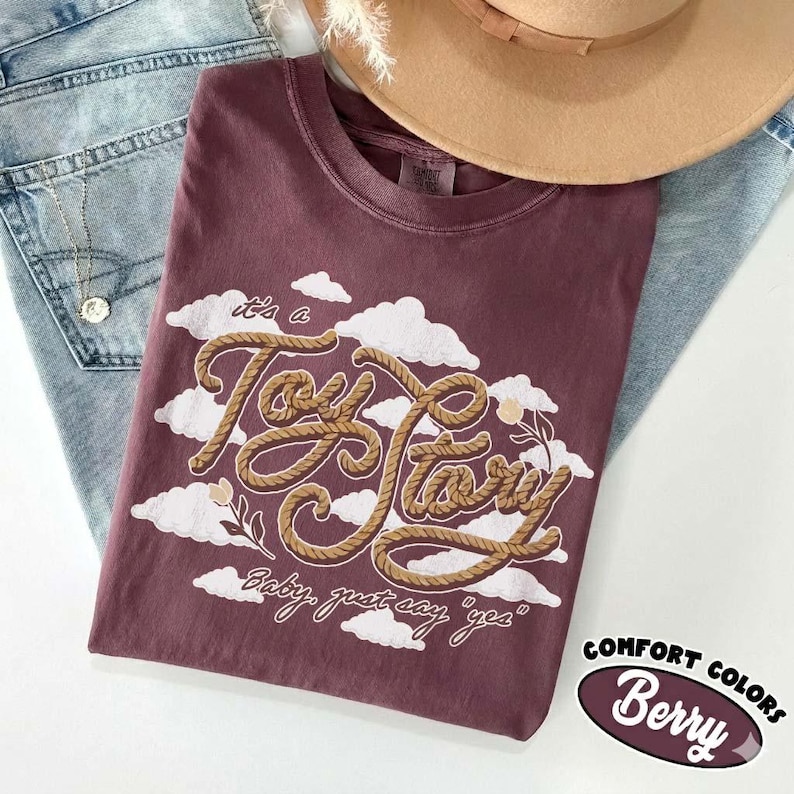 May include: A berry-colored t-shirt featuring a graphic of white clouds and the text "it's a Toy Story" written in a brown rope-style font, with the additional text "Baby, just say "yes"" printed below in a smaller white script font.