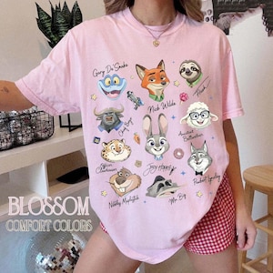 May include: A light pink t-shirt featuring cartoon character illustrations from Zootopia. The shirt includes the text "BLOSSOM COMFORT COLORS" and character names like "Nick Wilde" and "Judy Hopps".