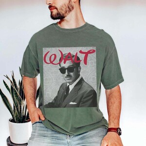 May include: Olive green t-shirt featuring a black and white image of a man wearing sunglasses, with the name "WALT" in red script above the image. The man is wearing a suit and tie.