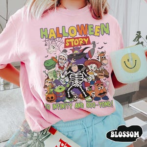 May include: Pale pink t-shirt with a Halloween-themed graphic featuring characters from a popular animated film. The graphic includes the text "HALLOWEEN STORY" and "TO INFINITY AND BOO-YOND!" A light blue mug with a smiley face is held in the image.