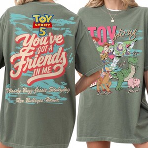 Pode incluir: Uma t-shirt verde-oliva com gr&aacute;ficos de Toy Story 5. As costas exibem o texto "You've Got A Friends In Me" com nomes de personagens. A frente mostra ilustra&ccedil;&otilde;es de Woody, Buzz Lightyear, Jessie, Bullseye, Rex e Hamm com o logo.