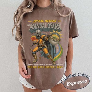 May include: A brown short-sleeve t-shirt featuring a graphic of The Mandalorian and Grogu. The print includes the text STAR WARS THE MANDALORIAN, BOUNTY HUNTER, and WHEN YOU CHOOSE TO WALK THE WAY OF THE MANDALORE YOU ARE BOTH HUNTER & PREY.