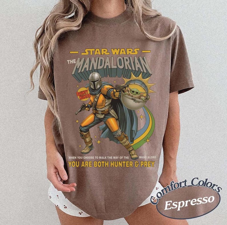 May include: A brown short-sleeve t-shirt featuring a graphic of The Mandalorian and Grogu. The print includes the text STAR WARS THE MANDALORIAN, BOUNTY HUNTER, and WHEN YOU CHOOSE TO WALK THE WAY OF THE MANDALORE YOU ARE BOTH HUNTER & PREY.